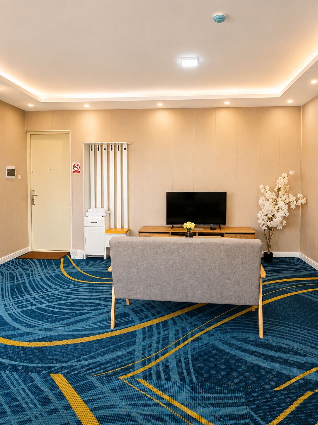 A hotel room with a flat-screen TV, a bench, a small table with a vase of flowers, a door with a no-smoking sign, and a patterned carpet.