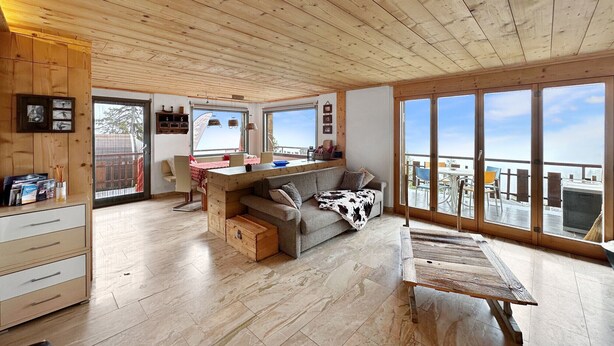 A spacious living area with a wooden ceiling, a sofa, a coffee table, and a sliding glass door leading to an outdoor balcony. - Mountain Bliss, view and Sauna - Swiss Alps (Anzère)