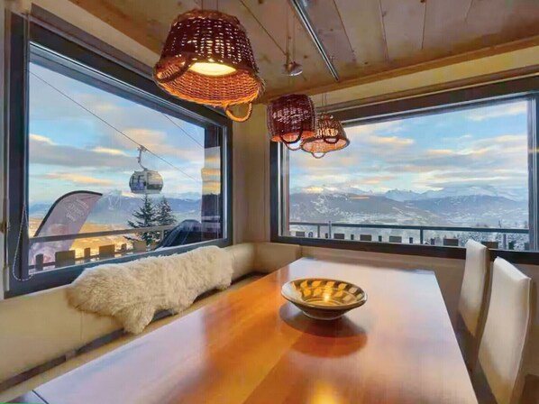 Ein Essbereich mit Holztisch, weiß gepolsterten Bänken und zwei hängenden Korblampen. Große Fenster bieten Blick auf eine Berglandschaft und eine Seilbahn.