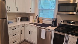 A modern kitchen with white cabinets, stainless steel appliances, a toaster, a coffee maker, and a window with blinds. - Downtown Marblehead 2brm/1.5bath w/ deck (Marblehead)