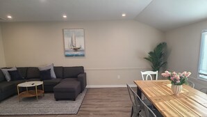 A living room with a sectional sofa, a coffee table, and a dining table with chairs. There is a painting on the wall and a potted plant on the right side. - Downtown Marblehead 2brm/1.5bath w/ deck (Marblehead)