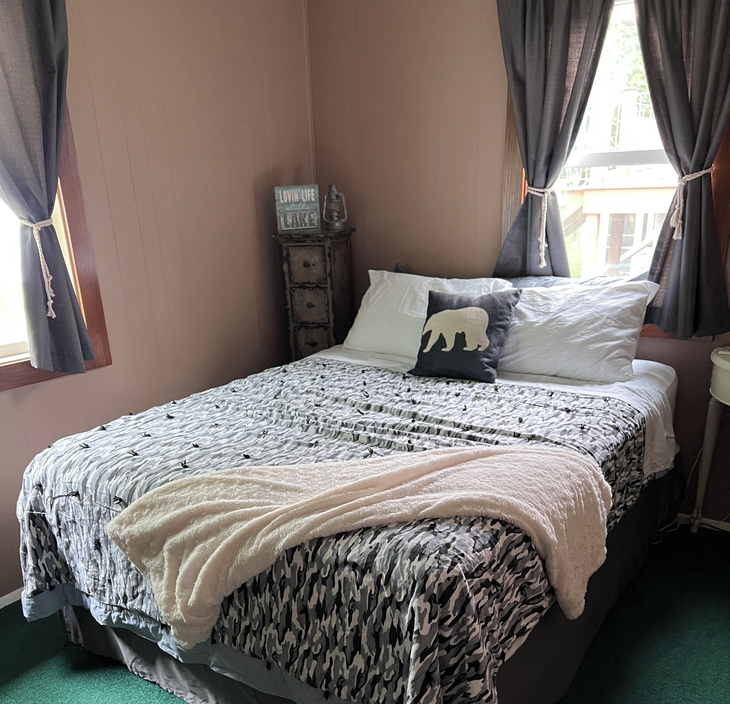 Ein ordentlich bezogenes Bett mit einem gemusterten Überwurf, einer weißen Tagesdecke und einem Kopfkissen mit einem Tier-Design. Ein kleiner Nachttisch mit einer Lampe und einem Schild mit der Aufschrift "Lovin' Life". Das Zimmer hat ein Fenster mit Vorhängen und Blick ins Freie.