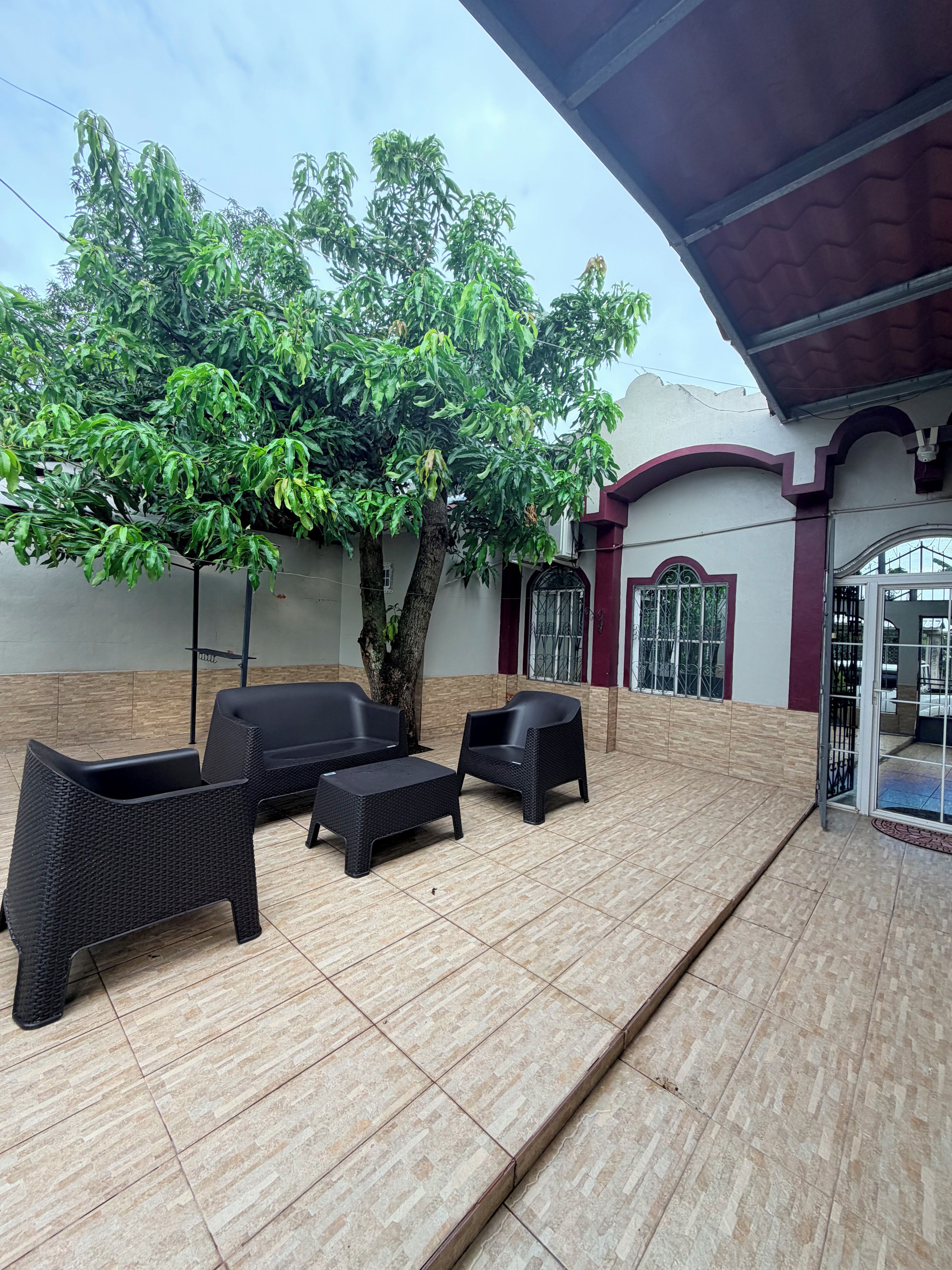 Un patio al aire libre con suelo de baldosas, mobiliario de mimbre negro y un gran árbol.