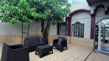 Un patio al aire libre con suelo de baldosas, mobiliario de mimbre negro y un gran árbol.