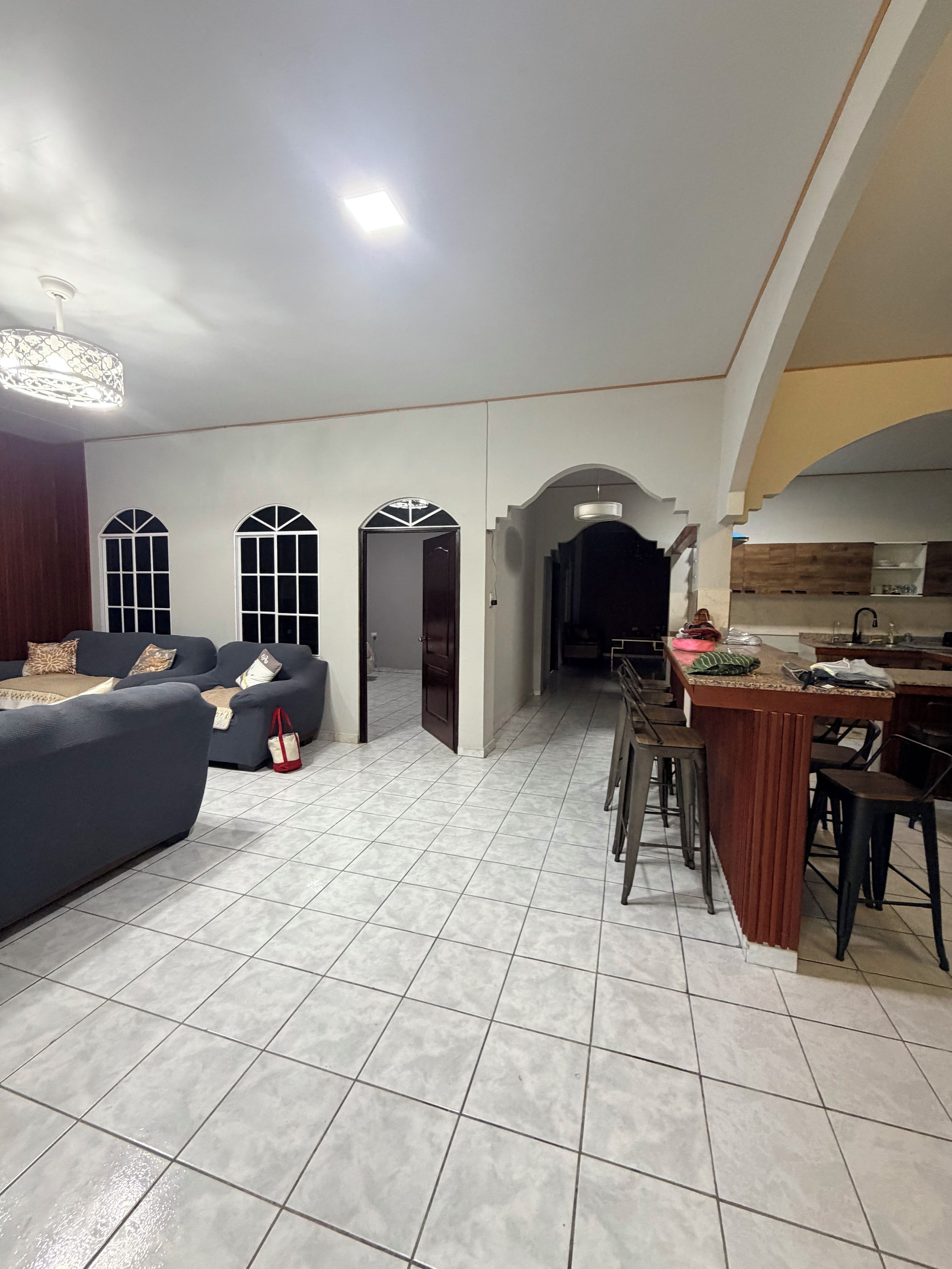Amplia sala de estar con un sofá de color oscuro, una cocina con barra y piso de baldosas.