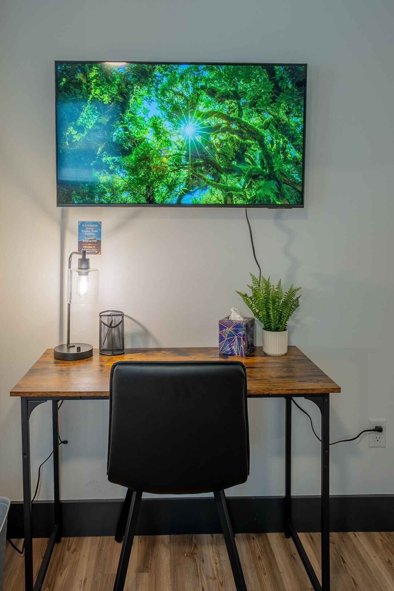 Uma mesa de madeira com uma cadeira preta, uma televisão fixada na parede, um abajur, uma caixa de lenços de papel e duas plantas em vasos.