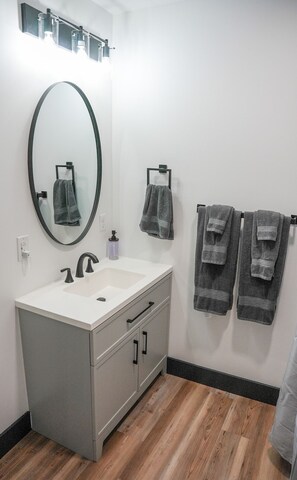 A modern bathroom with a large oval mirror, a white sink, and a vanity with drawers. There are two towels hanging on hooks and a bottle of soap on the sink.