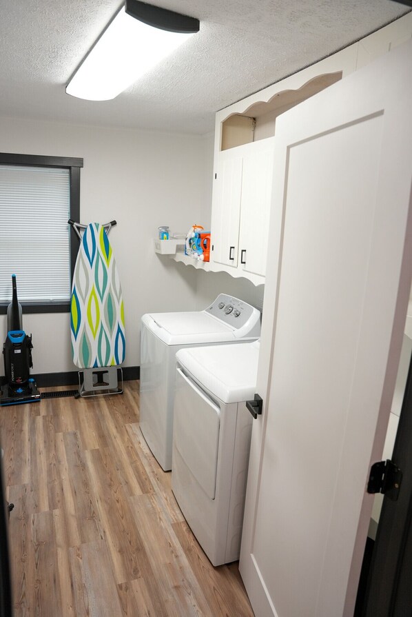 A laundry room with two front-loading washing machines, a vacuum cleaner, and a shelf with cleaning supplies.