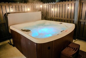 Un bain à remous avec une structure extérieure en bois et un intérieur blanc, entouré d’une clôture en bois et illuminé de guirlandes lumineuses.