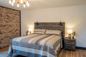 A bedroom with a wooden headboard, a striped bedspread, two bedside tables with lamps, and a brick wall.