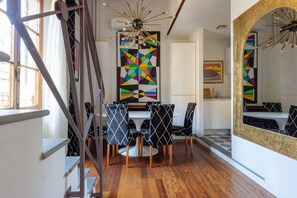 A dining area with a white table, black and white patterned chairs, a colorful geometric wall art, and a large mirror with a gold frame.