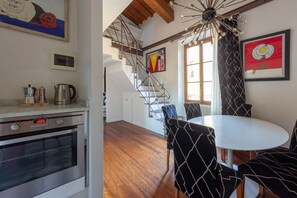 A modern kitchen with a built-in oven, a dining area with a white table and patterned chairs, a staircase, and artwork on the walls.