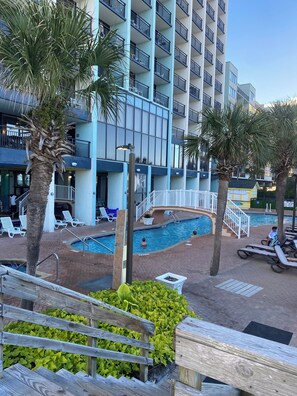 Zona de piscina de hotel con sillones de descanso, palmeras y un edificio con balcones.