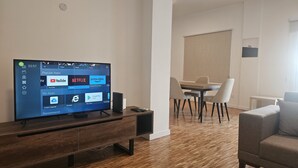 A modern living room with a flat-screen TV displaying a home screen with app icons, a wooden entertainment unit, a dining area with a table and chairs, and a grey sofa. - STAY Hydra Comfort Apartment (Nicosia)