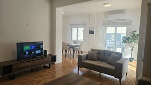 A modern living room with a flat-screen TV, a grey sofa, wooden flooring, a dining area with a table and chairs, and a potted plant. - STAY Hydra Comfort Apartment (Nicosia)