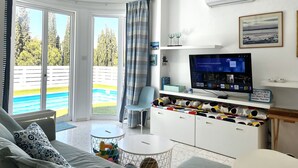 A modern living room with a large TV, a sofa, a dining table, and a view of the pool and trees outside. - STAY ALKYONI Villa (Protaras)