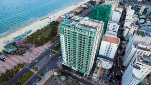 Un hotel moderno con un letrero llamativo que dice 'dalcorte', situado junto a una playa con palmeras visibles y una carretera con vehículos.