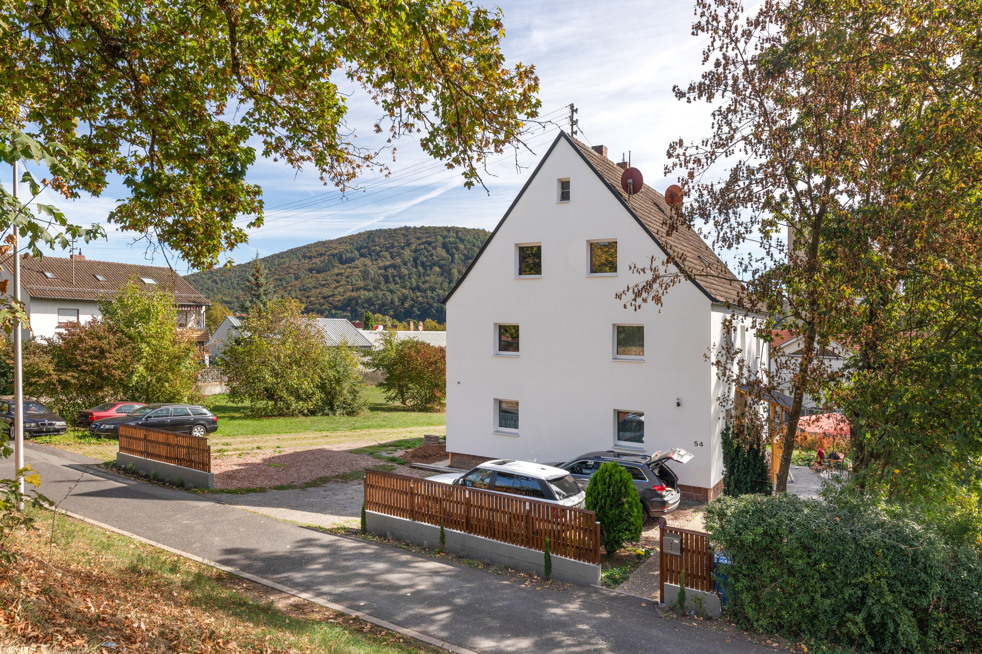 Une maison blanche à deux étages, avec un toit en pente, une allée bordée d’une clôture et des voitures garées.