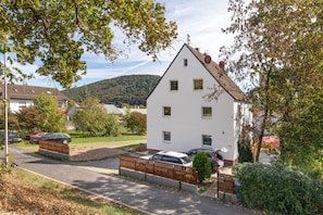 A white two-story house with a sloped roof, a fenced driveway, and parked cars. - Apartment 'Mirjam' (Lohr am Main)