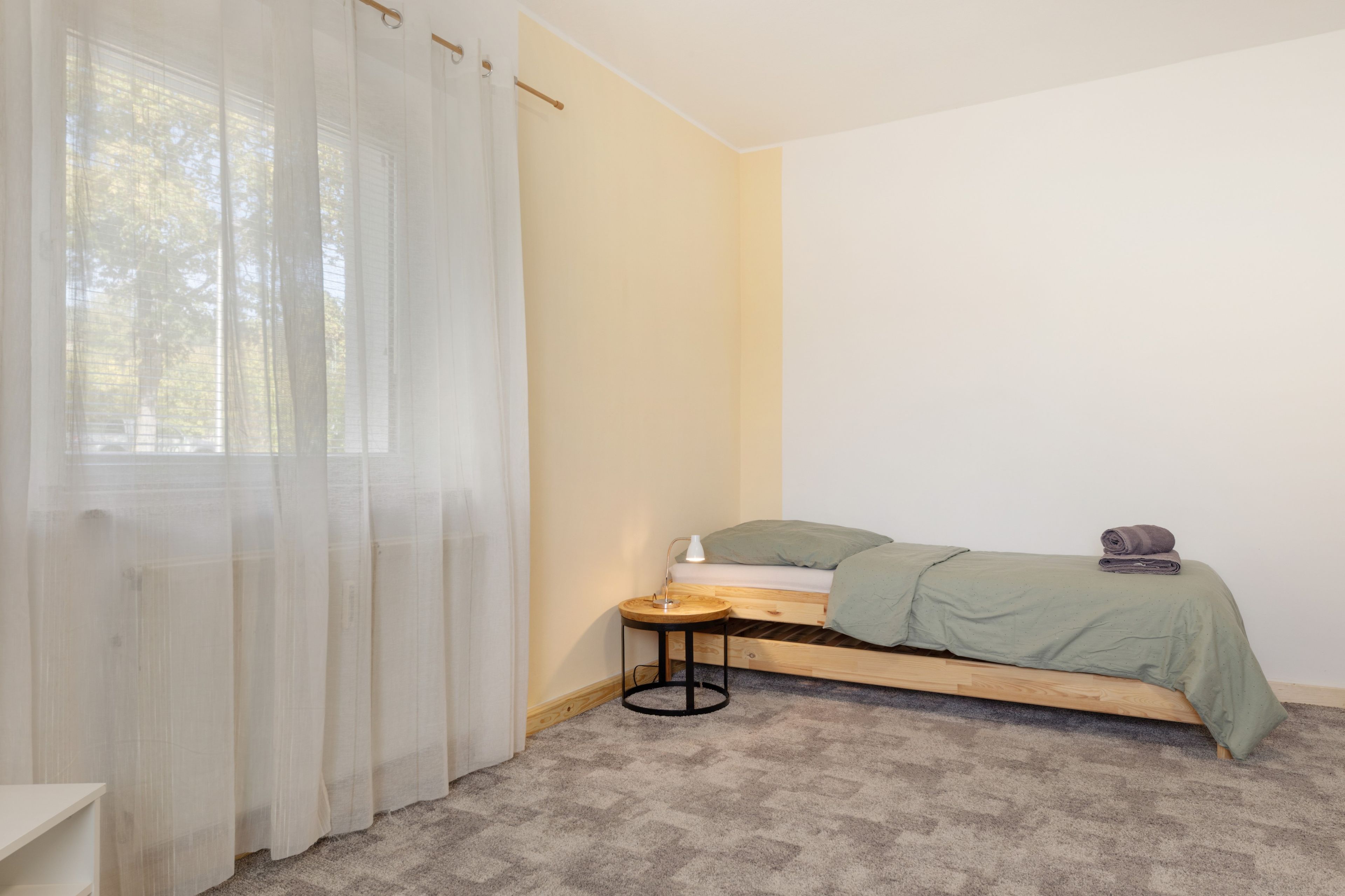 Een compact, verzorgd slaapkamer met een enkel bed, een nachtkastje en een raam met lichte gordijnen.