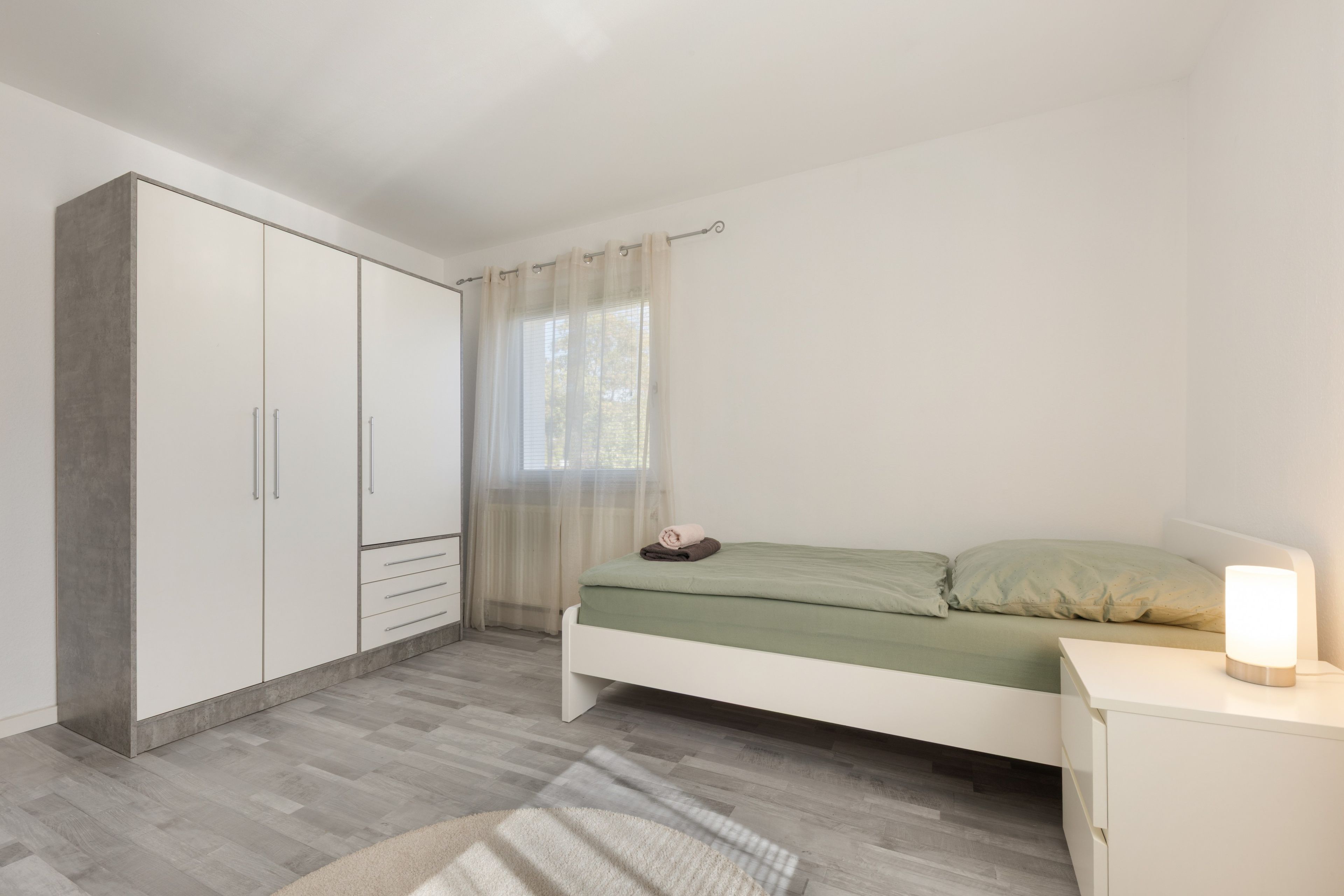 A bedroom with a bed, a bedside table with a lamp, a wardrobe, and a window with curtains.
