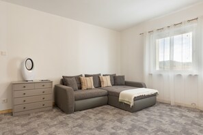 Een moderne woonkamer met een hoekbank, een bijzettafeltje en een raam met lichte gordijnen.