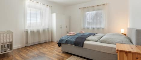 A bedroom with a bed, a crib, a wooden floor, and a window with curtains.