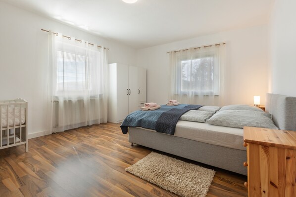 A bedroom with a bed, a crib, a wooden floor, and a window with curtains.