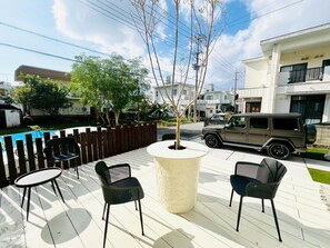 A patio with two black chairs and a central planter, a swimming pool, and a parked SUV. - Seaside Condo  Nagisa House (Chatan)
