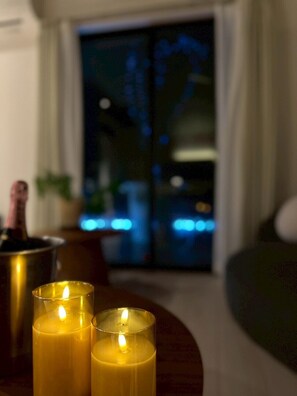 A living room with two lit candles on a table, a bottle of champagne, and a view of a cityscape at night through a glass door. - Seaside Condo  Nagisa House (Chatan)