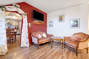 A living room with a sofa, a TV mounted on the wall, and a dining area visible through an open doorway.