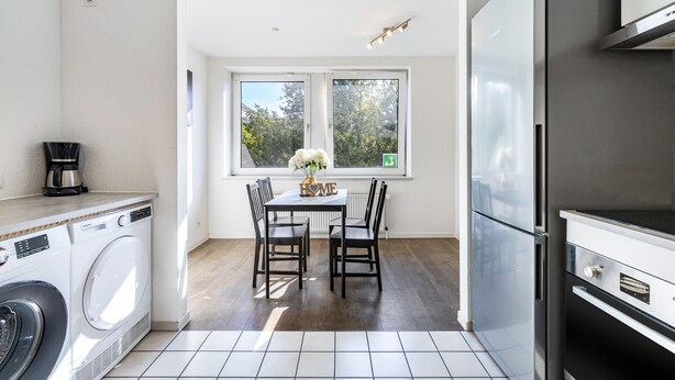 A compact kitchen with a washing machine, a dining table with chairs, a refrigerator, and a window with a view of trees. - Bright Duisburg Apartment for Business Stays & Old Town (Duisburg)