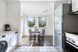 Cozinha compacta com máquina de lavar, mesa de jantar com cadeiras, geladeira e janela com vista para árvores.