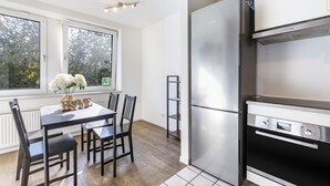 A modern kitchen with a stainless steel refrigerator, a dining table with chairs, and a window with a view of trees. - Bright Duisburg Apartment for Business Stays & Old Town (Duisburg)