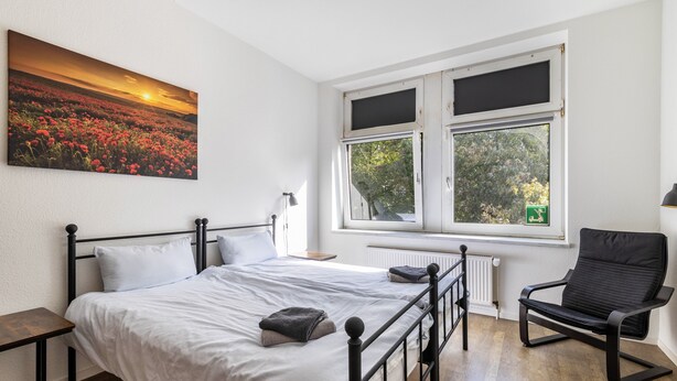 Un dormitorio con cama, una silla, una ventana y un cuadro en la pared.