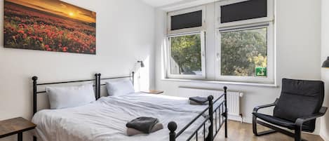 Un dormitorio con cama, una silla, una ventana y un cuadro en la pared.