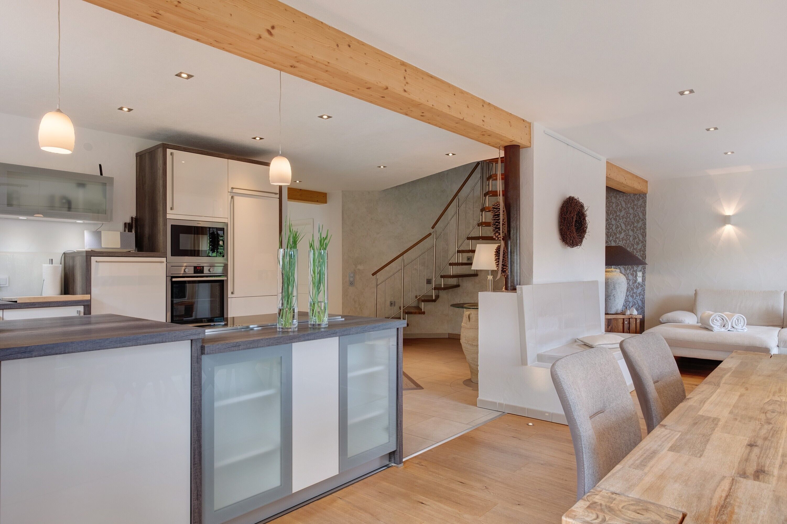 Una cocina moderna con una isla central, un comedor con mesa de madera, y una escalera que lleva a un dormitorio tipo loft.