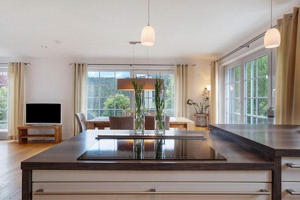A modern kitchen with a central island, wooden dining table, and chairs, large windows with curtains, and a flat-screen TV on a stand.
