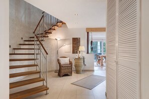 Una escalera con escalones de madera y barandillas de metal, una silla de mimbre con cojín, una lámpara de pie y un jarrón grande sobre una mesa.