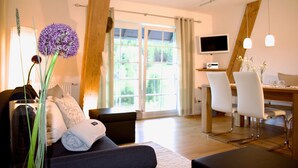 A modern living room with a sofa, a dining table, chairs, a TV, and a large window with a view of greenery. - Apartment '2 Natursuite' with Valley View, Shared Garden, Sauna and Wi-Fi (Furtwangen im Schwarzwald)