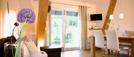A modern living room with a sofa, a dining table, chairs, a TV, and a large window with a view of greenery.