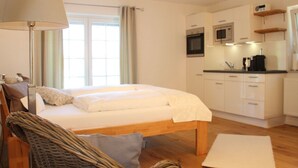 A bedroom with a bed, a bedside lamp, a wicker chair, and a view of a kitchen with cabinets and a microwave.