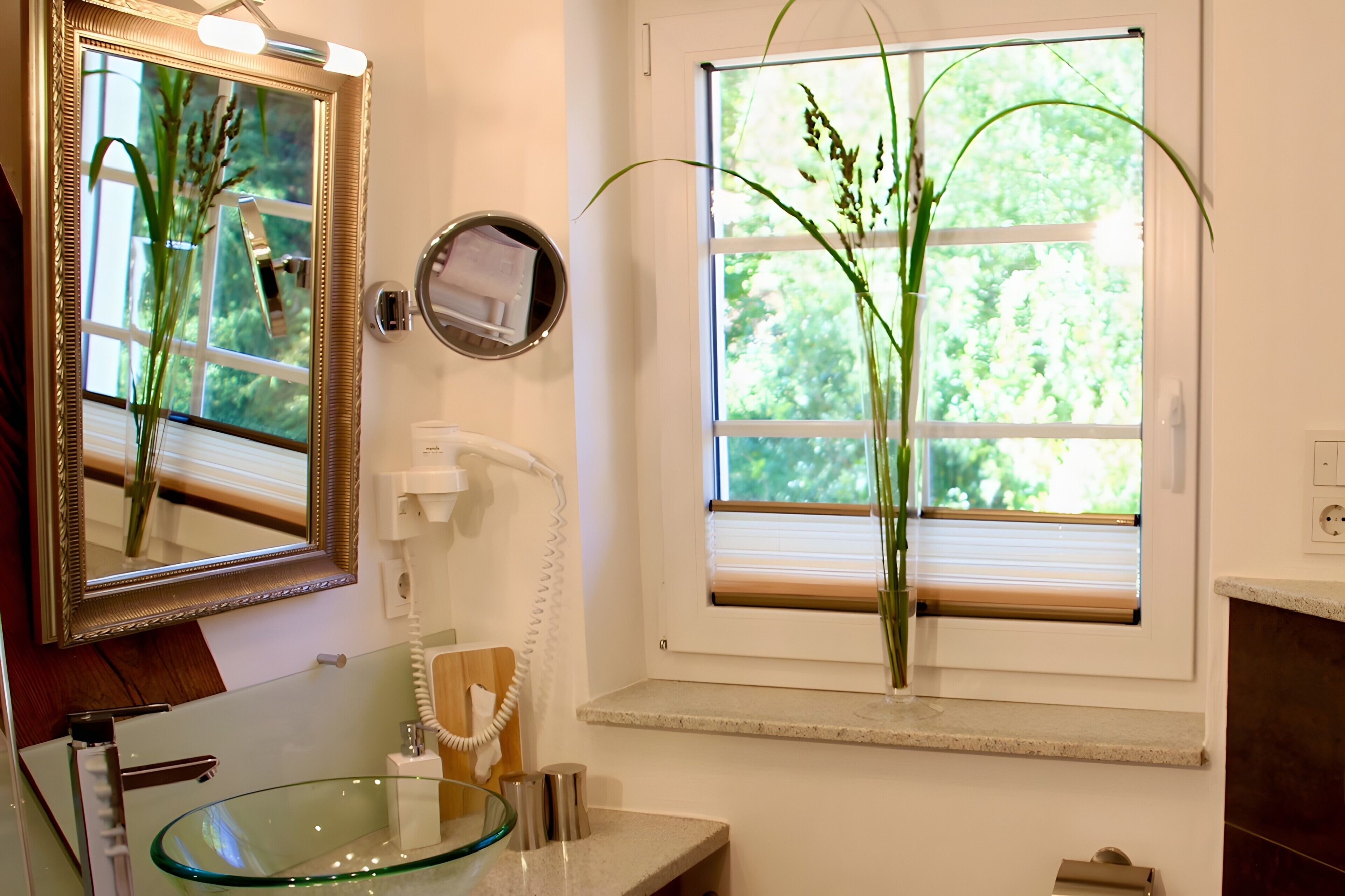 A bathroom with a glass sink, a mirror, a window with blinds, and a vase with plants.