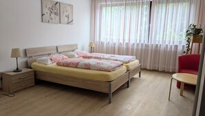 Ein Doppelbett mit floralem Bettzeug, ein Nachttisch mit Lampe, ein rotes Sessel, ein kleiner Tisch und ein Fenster mit Vorhängen.
