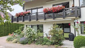 Un bâtiment à deux étages doté d’un balcon orné de fleurs rouges, d’un jardin bien entretenu avec différentes espèces de plantes et offrant une vue dégagée sur les environs.