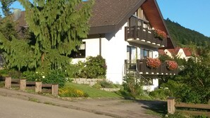Ein zweistöckiges Haus mit Balkon, umgeben von Grünpflanzen und einem Holzzaun.