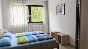 Una camera da letto con un letto, una finestra con tende, un termosifone, un comodino e un quadro appeso al muro.