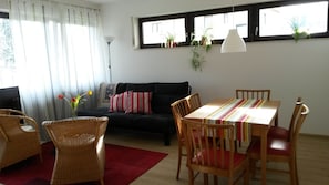Un salon avec un canapé, une table à manger et des chaises, un tapis rouge et une fenêtre ornée de plantes.
