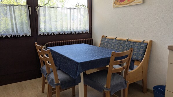 A dining area with a table covered by a blue tablecloth, wooden chairs with blue cushions, a radiator, and a window with lace curtains.