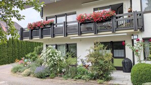 Ein zweistöckiges Gebäude mit einem Balkon, der mit roten Blumen geschmückt ist, ein Garten mit verschiedenen Pflanzen und ein Stuhl davor.
